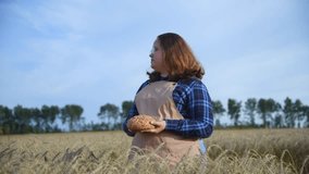 Portrait of cheerful brown haired plus size caucasian woman in eyeglasses wearing blue checkered shirt and beige apron holding loaf of bread on agricultural field in a summer day. Real time video.