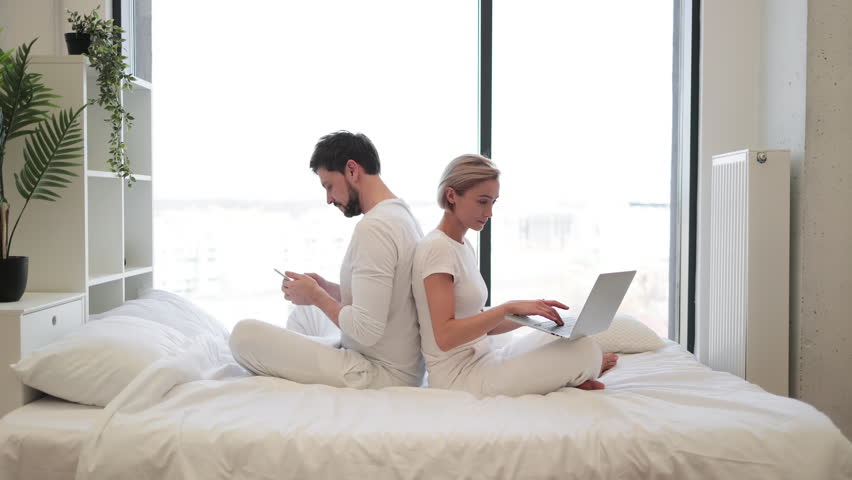 Cheerful amorous people sitting back to back while spending time together in spacious apartment at weekend. Joyful caucasian family scrolling social media on wireless gadgets.