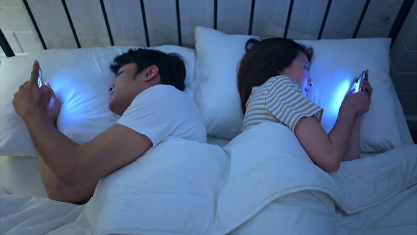 Young couple sleeping on the bed in the bedroom at night time with their backs to each other and playing on their phones, ignoring each other.