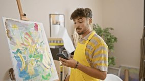 Handsome young arab man artist, happily learning to draw on canvas at a vibrant art studio, immersed in a confident video call lesson