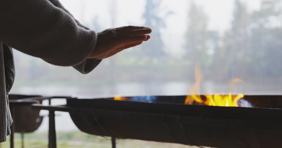 Hands, camping and fire with a person in the forest, getting warm by a bbq for heat while outdoor in the wilderness. Hiking, travel and an adult in nature to camp with a burning flame for survival