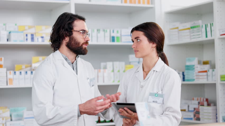 Pharmacists discussing stock orders and medication treatment while working on a tablet in a pharmacy. Two healthcare professionals discussing a patient prescription in a dispensary