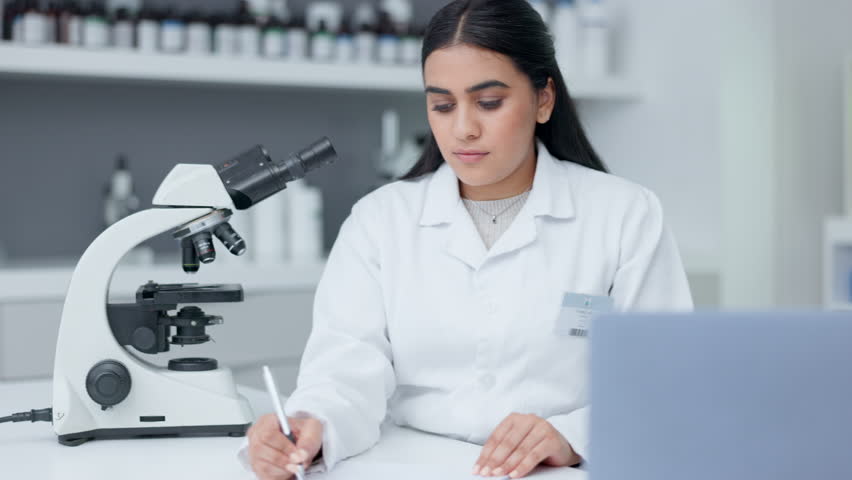 Laboratory scientist looking into a microscope to examine a sample on a slide. Biochemist engineer searching for breakthrough virus cure through medical research and writing discovery on clipboard