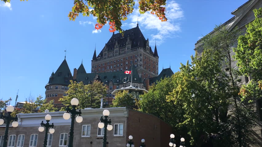Chateau Frontenac seen from below with antique streetlights with globe lights. Pan in to flag of Canada waving in the wind. Day, blue sky.