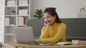 Yellow sweater woman is typing and greeting friends via video call on laptop with happy face.