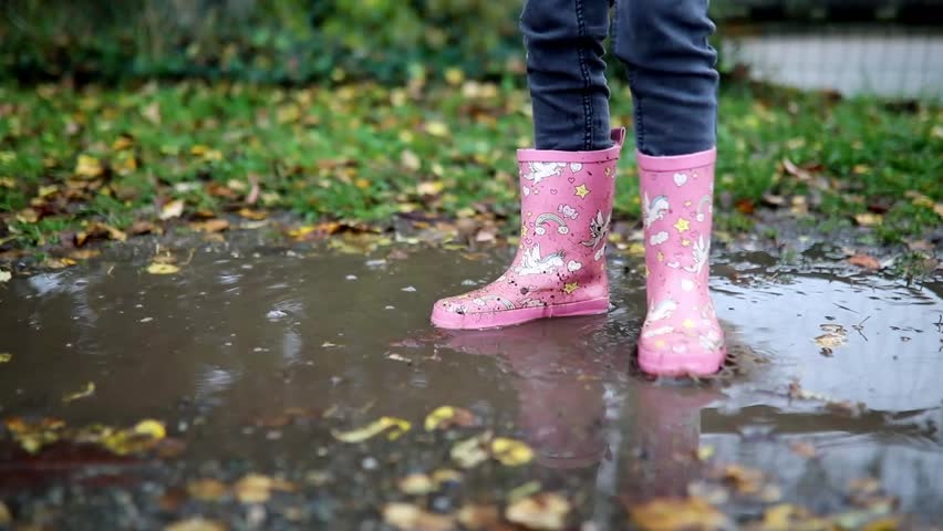 Child wearing pink rain boots with unicorns and jumping in puddle on a fall day
