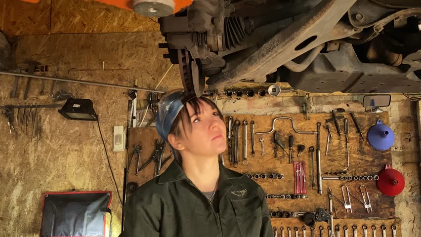 Young woman in coveralls working at a car service station, performs technical car maintenance and repairs the vehicle