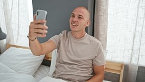 Young hispanic man doing video call with smartphone sitting on the bed at bedroom