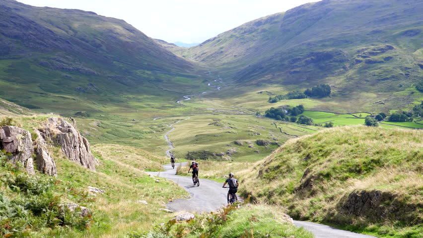 Rear view of bicycle rider group of four riding downhill mountain road towards Duddon Valley in Lake District, England, United Kingdom, one cyclist stops to take a picture, Hard Knott Pass viewpoint.