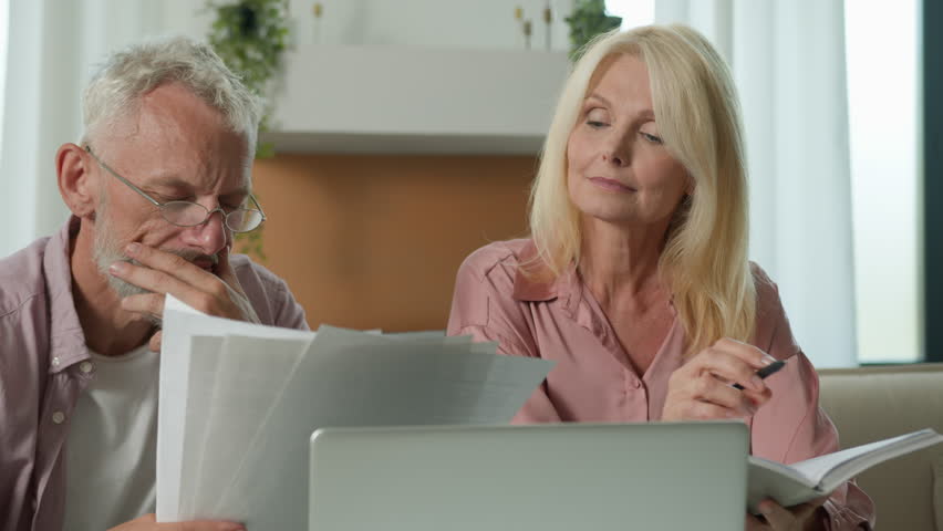 Focused pensive elderly retired married couple holding paper documents checking analyzing calculating bills bank loan payment doing paperwork unpaid debt taxes make online payment manage family budget Royalty-Free Stock Footage #1110966927
