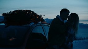 Silhouette of a couple kissing near the car with lit Christmas tree tied to the roof, winter mountains in the background - Powered by Shutterstock - Get 15% off with code: PIKWIZARD15