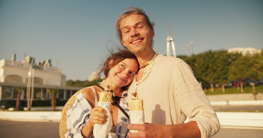 Happy couple portrait guy and girl eating hot dog on modern beach in the morning. Blond guy in light clothes III brown-haired girl goes hot dogs on a sunny beach in the morning
