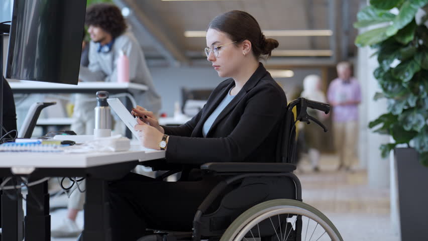 A businesswoman in a wheelchair is seen working on a laptop in a modern office, exemplifying a workplace that values inclusivity, accessibility, and professionalism. Royalty-Free Stock Footage #1110974009
