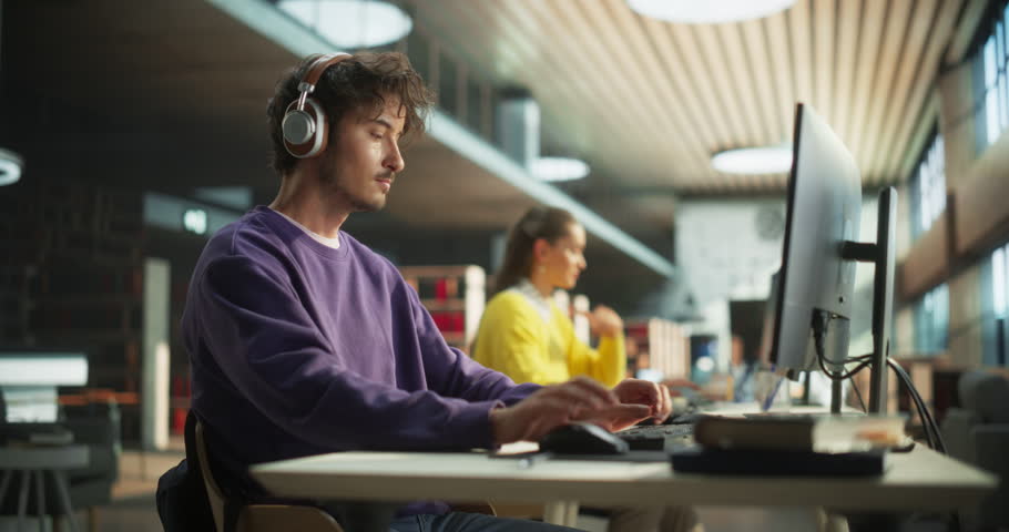 Portrait of a Stylish Male Student Working on His University Project on a Desktop Computer. Young Handsome Man Studying a Course Online in a Quiet Public Library with Bookshelves with Books