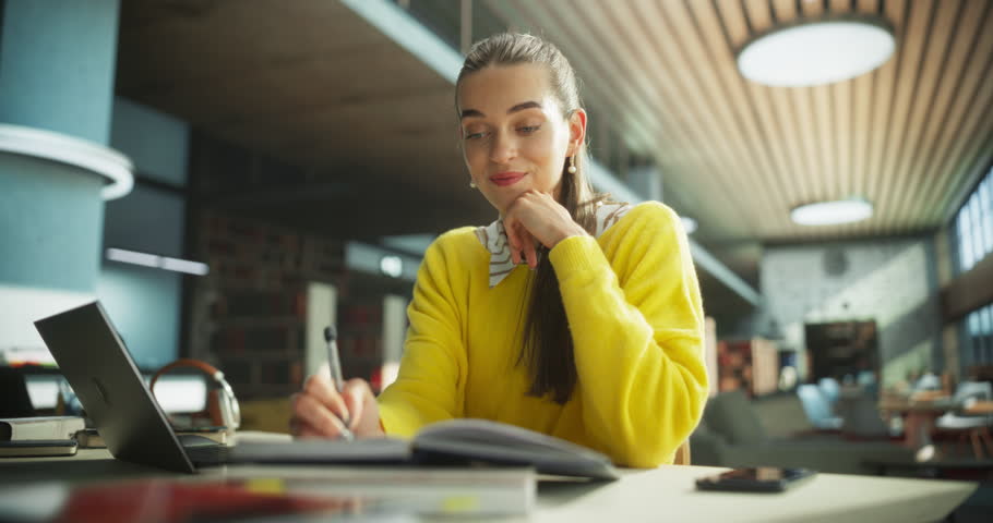 Happy and Focused Student Studying in a Modern Library. Young Female Enjoying Time Alone for Thinking and Problem Solving. Girl Using Laptop Computer to Work on a University Research Project Online