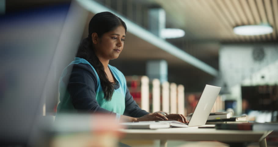 Focused Indian Female Working on a College Project for Class Assignment, Using Online Office Software on Laptop Computer in a Public Library Space. Young South Asian Woman Learning New Skills Online Royalty-Free Stock Footage #1110977499
