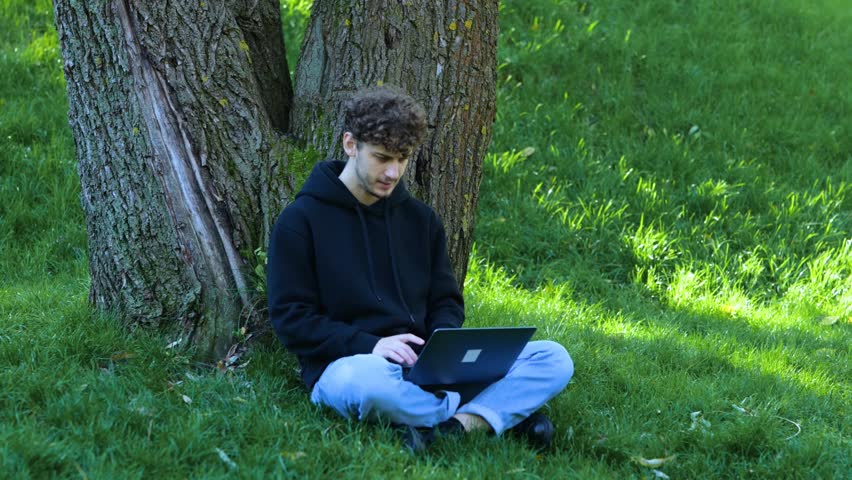 Young man sitting in the park, leaning against a tree while working with laptop and typing. Autumn season. Education, working concept. Real time concept.