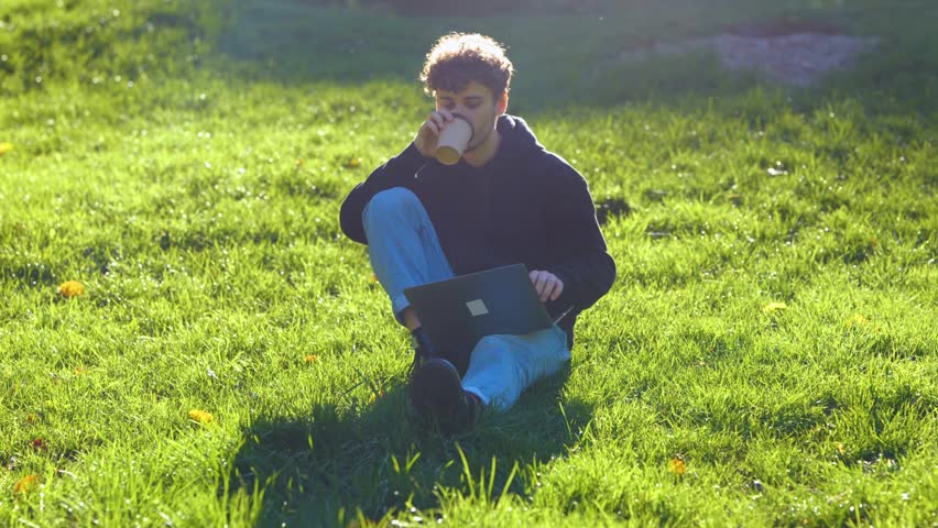 Young man working with laptop and drinking coffee. Guy sitting on the grass in the park. Autumn season. Education, working concept. Real time concept.