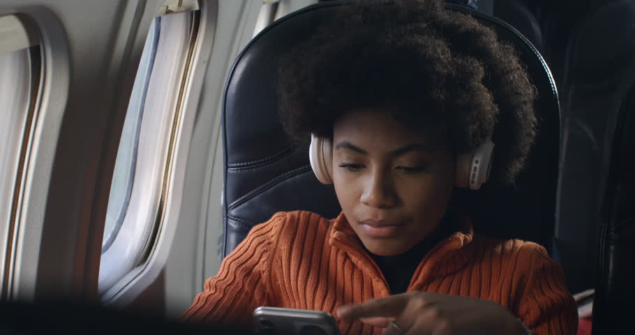 Close up of young african woman with headphones listening her favorite music via smart mobile phone and looking outside window of aircraft while flying at airplane to holidays vacation destination. Royalty-Free Stock Footage #1110983795