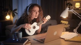 Pretty young curly woman watch video tutorial on ukulele guitar playing or having personal lesson with teacher distance remote on laptop computer at evening at home Online musical education concept