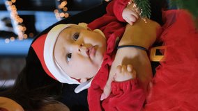 Vertical video. Cute baby in Santa costume looking at Xmas tree, mother holding her adorable infant daughter in hands at home. Winter holidays and New Year concept
