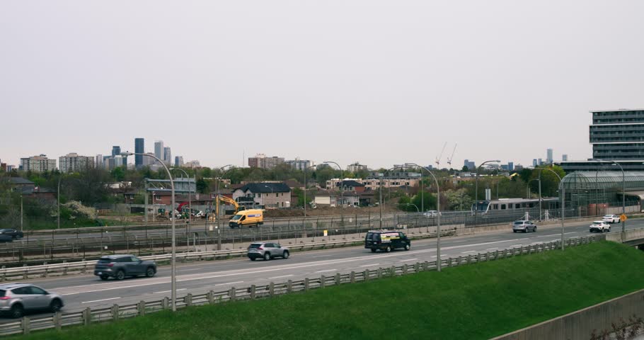 Subway train travels from city on railway along residential buildings. Wide view of urban cityscape with train, highway with rushing cars, houses, greenery grass. Concept use train to come on time