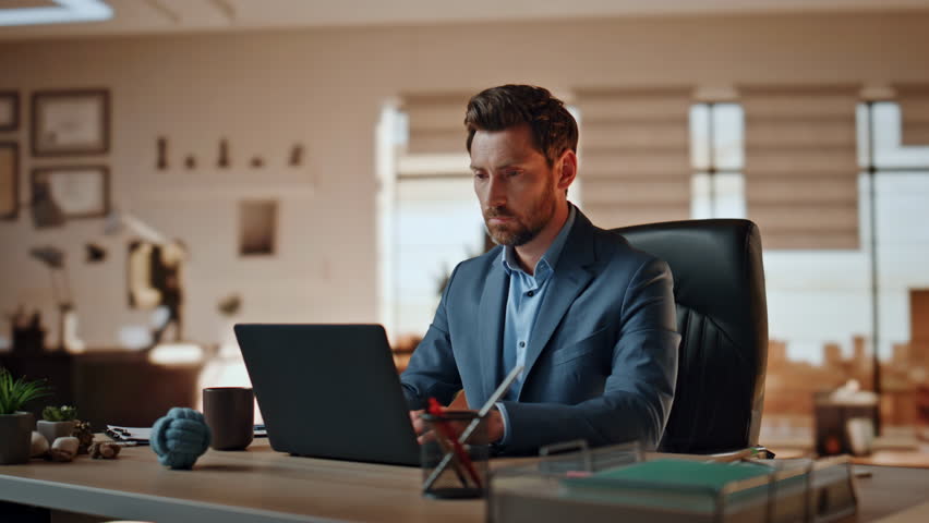 Smiling businessman answering call at laptop table workplace. Manager watching computer talking at panoramic window office. Happy boss gesturing hand reacting great news at mobile phone conversation 