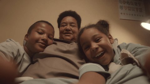 Handheld POV of cheerful African American tween kids recording video of themselves and their happy grandma having fun together in bed at nightの動画素材