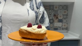 4K video. Woman eats cheesecake with raspberries with fork in modern European kitchen. Female hand holds orange plate with fresh sweet delicious cake close-up. Concept of healthy food, pastry shop	