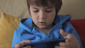 Child Playing Games In Phone at Home Sitting on Couch. Boy Playing Video Game on Mobile Phone. Preschooler Plays Video Game Smartphone on Sofa. Kid Using Phone for Gaming Online Education Social Media