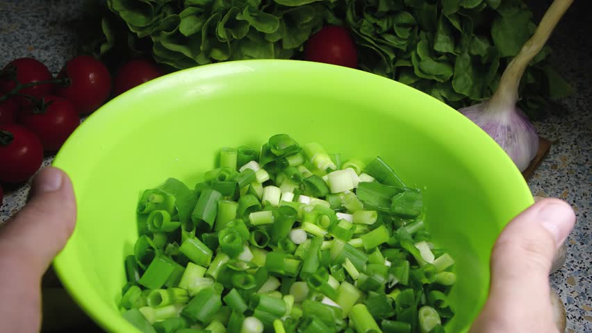 Hands toss pieces of chopped green onions in a plastic bowl. Different vegetables laid at foreground. 200 fps slow motion.