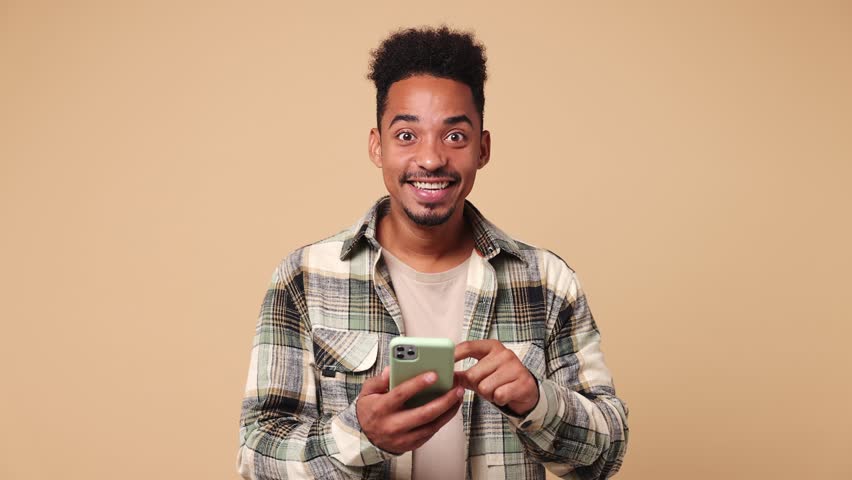 Happy joyful young man of African American ethnicity he wear shirt casual clothes hold use point finger on mobile cell phone just found out great big win news isolated on plain beige brown background