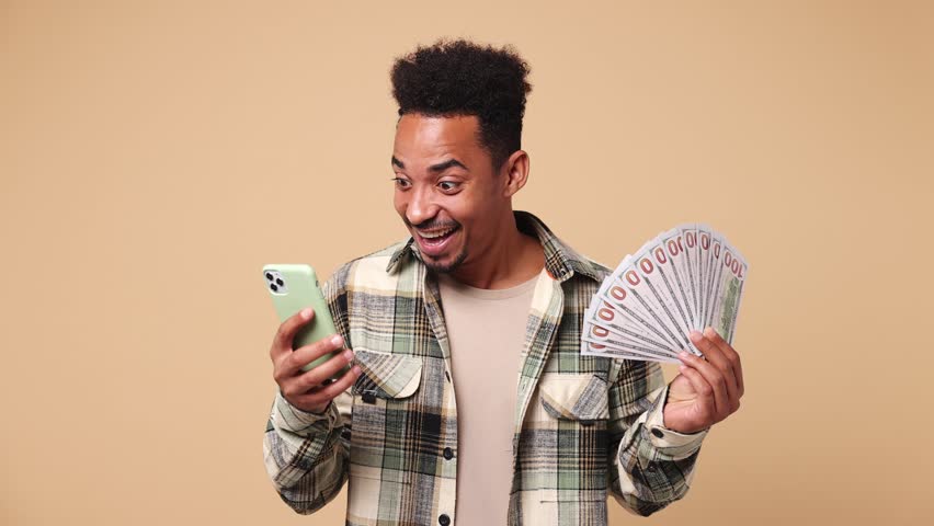 Young joyful excited overjoyed man of African American ethnicity wear shirt casual clothes use mobile cell phone hold win fan of cash money in dollar banknotes isolated on plain beige brown background
