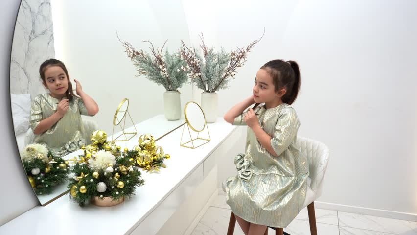 Fashion cute girl kid in beautiful dress sitting at the makeup table looking into a large mirror and straightening her hair. Pretty little lady admires her reflection in the mirror.