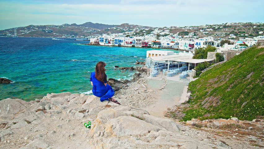 A beautiful woman in a blue dress admiring the view of the crystal blue sea with whitewashed houses in Mykonos. Fashionable girl with long hair enjoying a cinematic view of the Mykonos landscape.