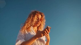 Woman using mobile phone and smiling. Blonde lady with curly hair looking on screen of cellphone. Young woman using smartphone app on blue sky background. Low angle view - Powered by Shutterstock - Get 15% off with code: PIKWIZARD15
