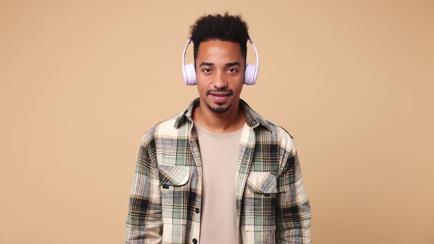 Young man of African American ethnicity he wears shirt casual clothes listen mp3 music in headphones slow dance sing song have fun enjoy relax show thumb up isolated on plain beige brown background | Shutterstock HD Video #1111038459