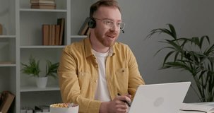 Casually dressed man with eyeglasses has video call with his business partners. Man with ginger beard using headphones while speaking with office colleagues. Bowl with colorful cereals on desk