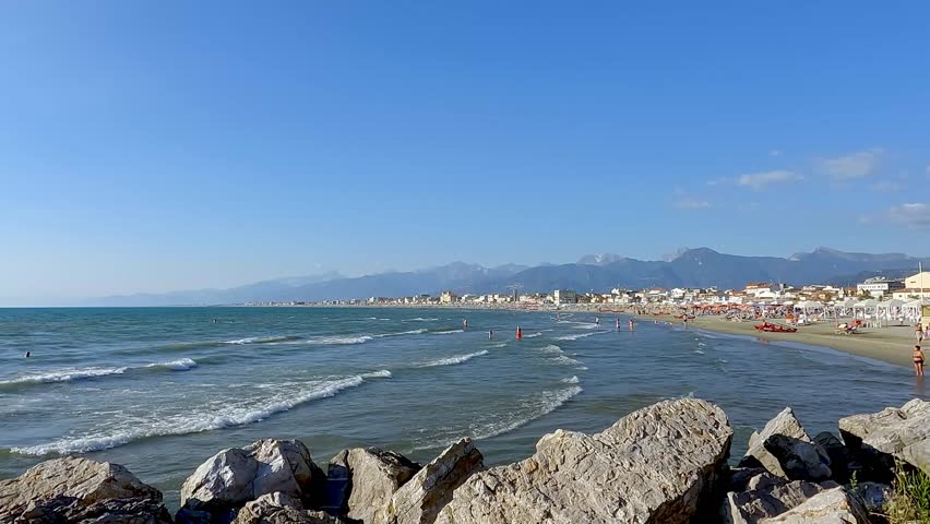 A sunny day on the beach in Viareggio, Tuscany, Italy with small waves. Parasols and sun beds along the beach. Some people are in the water. Mountains in the background