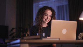 Charming young curly woman sitting at desk and flirting talking on laptop by video chat conference meeting with boyfriend at late night Remote distance relationships concept 