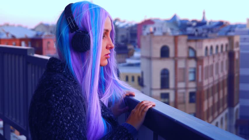 A white girl with long colored hair in dark headphones in a dark coat looks from the balcony at the city 4K 24 fps