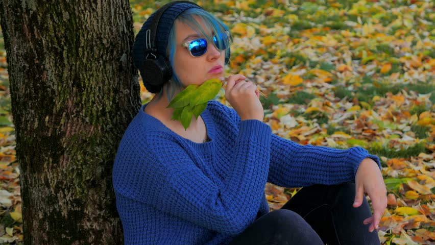 A white girl with colored hair in sunglasses, dark headphones, a blue sweater against a background of bright yellow autumn leaves 4K 24 frames