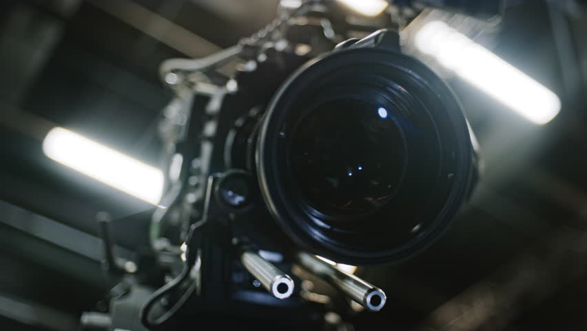 Low angle shot of a professional cinema camera and long telephoto zoom lens, zoom motor is turning the gears and the lens element is moving to change focal length. Studio ceiling in background.