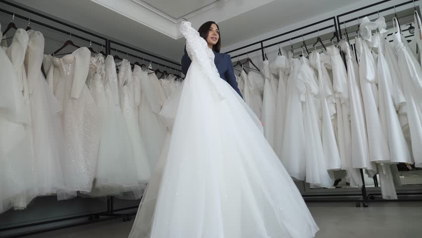 A happy young bride in a wedding shop. A woman is holding a wedding dress on a hanger in her hands, she has put the dress to her body and is spinning. Fitting a wedding dress. Royalty-Free Stock Footage #1111065091