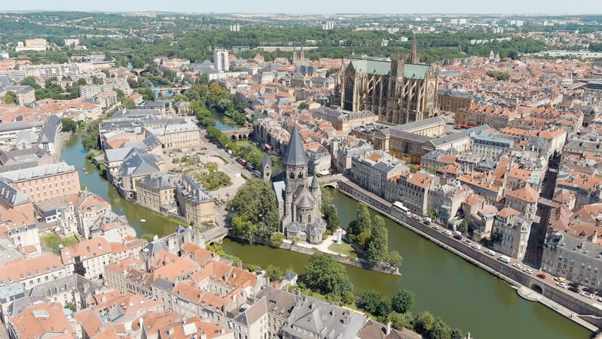 Metz, France. Temple Neuf, a Protestant church built in Romanesque Revival style from grey sandstone, stands on the Ile du Petit-Saulcy surrounded by the Moselle River. The church was inaugurated in 1904 in the presence of Emperor Wilhelm II and has been a French monument historique since 1930. Its dark stone mass contrasts with the lighter limestone buildings of the surrounding historic city center. The camera captures a smooth aerial forward movement over the river, revealing the church