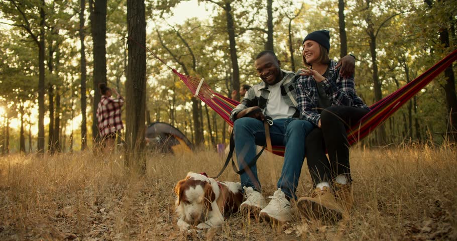 A happy couple, a man with Black skin and a girl in a black hat in a plaid shirt, are sitting on a Red hammock, next to them is their white-brown dog, and behind them is the rest of the group of Royalty-Free Stock Footage #1111070177