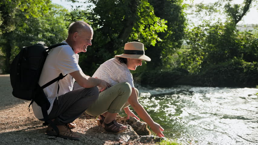 fun in retirement, joyful elderly married couple having fun splashing water while sitting by river in park on a sunny day