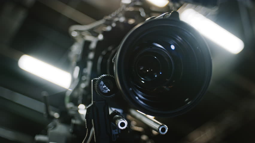 Low angle shot of a professional cinema camera and long telephoto zoom lens, focus motor is turning the gears and the lens element is moving to change focus distance. Studio ceiling in background.