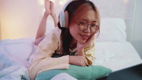 Positive young asian woman in eyeglasses watching video clip on laptop with headphones and singing on the bed at home