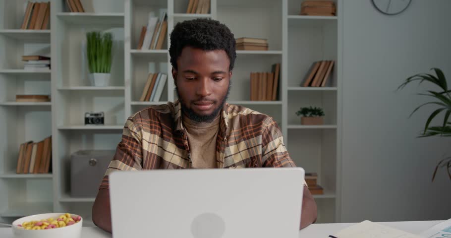 Portrait of african man working in the stylish office, looking at camera. Casually dressed manager sitting at the desk, using laptop. Bowl with colorful cereal for lunch standing on the desk. | Shutterstock HD Video #1111086817